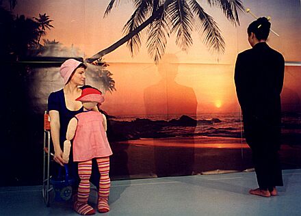 'Die Tanzstunde' (Probenaufnahme): Elisabeth Breckner mit Puppe; rechts wirft Mari Honda ihren Schatten auf die projizierte Strandkulisse.